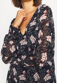 Blumenbluse mit langen, gerafften Ärmeln, tiefem V-Ausschnitt, dunklem Hintergrund, rosa-weißen Blumenmuster und leichtem Stoff.