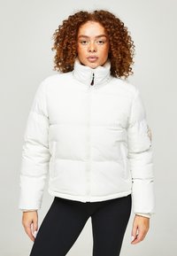 Witte pufferjack met een hoge kraag, een rits aan de voorkant en zichtbare stiksels. Beschikt over een textuurpatch op de linker mouw. Zachte afwerking.