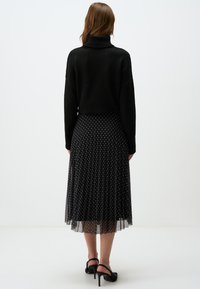 Maglione nero a collo alto abbinato a una gonna nera a pieghe con motivo a pois bianchi. La gonna presenta un sovrapposto trasparente. Sono visibili tacchi neri.