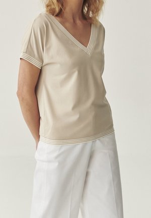 Femme portant un haut beige à manches courtes avec col en V et bordure rayée, et un pantalon blanc ample, debout devant un fond clair uni.