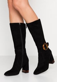 Botas negras de ante hasta la rodilla con cremallera en el lado, tacón grueso y un acento de carey circular en el tobillo.