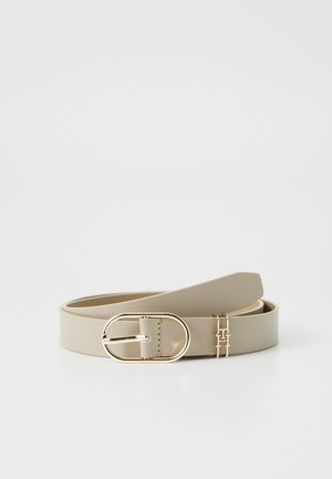 Ceinture en cuir beige avec une boucle ovale dorée et un petit pendentif décoratif doré sur un fond clair uni.