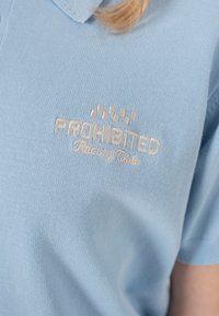 Chemise en tricot bleu clair avec texte brodé beige "PROHIBITED Racing Club" et un petit drapeau à damier au-dessus du texte.
