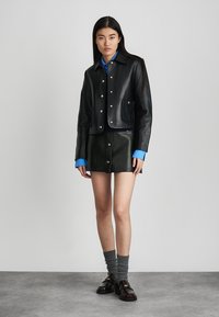 Giacca di pelle nera con bottoni a pressione argentati, tasca frontale, indossata sopra una camicia blu. Abbinata a una minigonna di pelle nera e calze grigie.