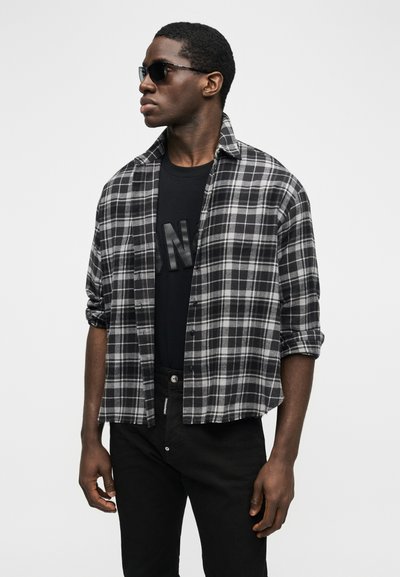 Νεαρός άνδρας που φοράει μαύρα γυαλιά ηλίου, μαύρο μπλουζάκι με σχέδιο, από πάνω ένα ασπρόμαυρο καρό πουκάμισο και μαύρο παντελόνι.