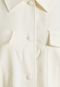 Lähikuva valkoisesta kangastakista, jossa on kaulus, neljä pyöreää peitettyä nappia ja kaksi läpällistä rintataskua.