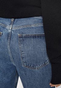 Ausgewaschene blaue Jeans mit mittelhohem Bund, ausgestattet mit einer Gesäßtasche und sichtbaren Nähdetails. Kombiniert mit einem schwarzen Strickoberteil.