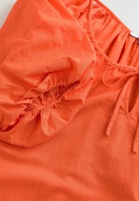 Pomarańczowa bluzka z marszczonymi rękawami, dekoltem ze sznurkiem i lekką, teksturowaną tkaniną. Gładka powierzchnia z lekkim połyskiem.