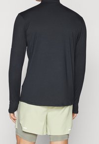 Schwarzes Langarmshirt mit hochgeschlossenem Kragen, glatter Textur und leicht taillierter Passform, kombiniert mit hellgrünen Shorts mit seitlichen Schlitzen.