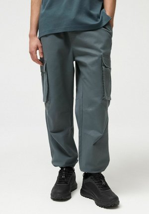 Osoba w turkusowych spodniach typu cargo z bocznymi kieszeniami i czarnych sportowych butach na sznurowadła, stojąca na tle jednolitego, jasnego tła.