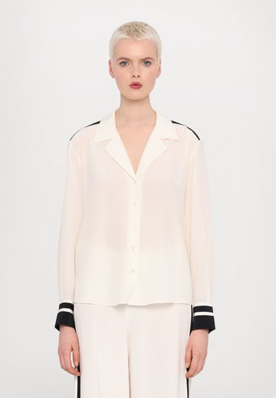Blouse de couleur crème en matière transparente, avec un col à revers et des poignets à rayures noires, dotée d'une fermeture à boutons et d'une coupe décontractée.