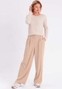 Beige strukturierter Pullover mit langen Ärmeln, kombiniert mit weiten beigen Hosen, die über vordere Falten und einen Knopfverschluss verfügen. Weiße Sneakers.