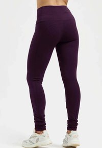 Lila leggings med hög midja, figurnära design, slät textur och en rynkad avslutning vid anklarna. Matchas med vita träningsskor.