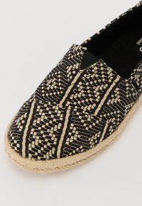 Svart og beige vevet slip-on sko med rund tå, med flettet jutekant og elastiske sidepaneler for enkel påkledning.