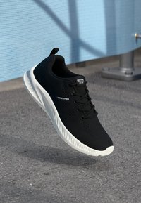 Sort atletisk sko med et tekstureret stofoverdel, hvid sål og snørebåndsdesign. Har branding på siden og en løkkebånd bagpå.