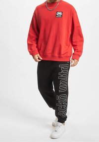 Rode sweatshirt met een logo patch, gecombineerd met zwarte sweatpants met grijze tekst. Witte sneakers maken de sportieve look compleet.