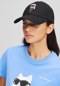 Svart bomullslue med et grafisk motiv prydet med rhinestones foran. Enkelt svart design med buet skjerm.