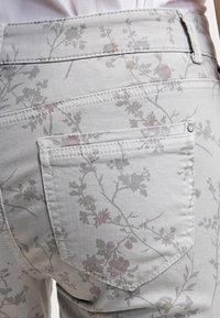 Grå denimjeans med ett blommönster i dämpade toner och en fickdetalj. Slät textur med kontrasterande sömmar längs sömmarna.