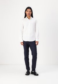 Camisa blanca de manga larga con botonera, combinada con pantalones oscuros de color marino y zapatos negros, sobre un fondo simple.