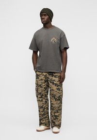 Νέος άνδρας που φοράει σκούφο σε σκούρο χρώμα, ξεβαμμένο γκρι T-shirt με σήμα, παντελόνι καμουφλάζ και άσπρα αθλητικά παπούτσια, στέκεται μπροστά σε ένα απλό φόντο.