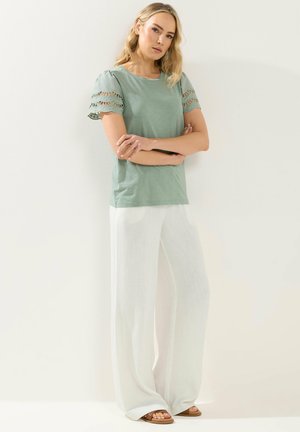 Kvinde med blondt hår står op ad en hvid væg iført en salviegrøn bluse med korte ærmer og udskæringer på ærmerne samt vide hvide bukser og sandaler.