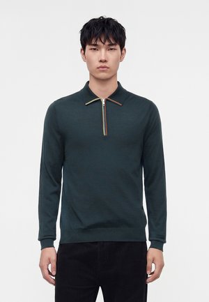 Jeune homme portant un polo à manches longues vert foncé avec un détail de fermeture éclair rayé multicolore, debout devant un fond clair uni.