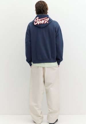 Person trägt einen marineblauen Hoodie mit rot-weißem grafischem Text auf der Kapuze, beige weite Hosen und ein gestreiftes Hemd darunter, von hinten gesehen.
