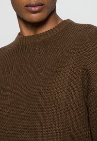 Brun stickad tröja med ett texturerat mönster, rundhalsad och ribbade ärmslut. Materialet verkar mjukt och varmt.