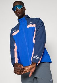 Giacca a vento blu e blu navy con dettagli in rosa e logo, dotata di chiusura con zip e polsini elastici. Indossati occhiali da sole riflettenti.