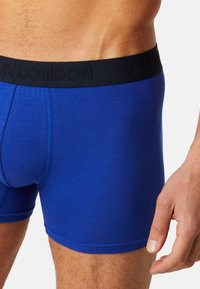 Blue cotton boxer shorts with a black elastic waistband featuring embossed lettering. Smooth texture and fitted design.