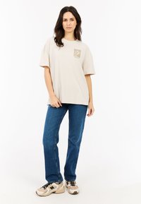 Beige katoenen t-shirt met korte mouwen en een vierkante zak met een geprint ontwerp. Gecombineerd met blauwe jeans met rechte pijpen en sneakers.