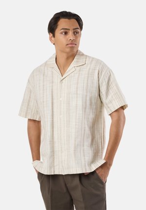 Young man wearing a beige patterned short-sleeve button-up shirt and dark pants, standing with hands in pockets, looking to the side.