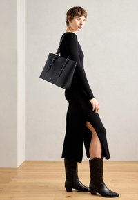 Czarna sukienka z żebrowanym wzorem i wysokim rozcięciem na udzie, w połączeniu z wysokimi czarnymi skórzanymi botkami oraz usztywnioną czarną torbą tote z kontrastowym przeszyciem.