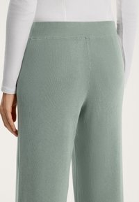 Světle zelené žebrované pletené kalhoty s vysokým pasem, které mají hladkou texturu a design s rozšířenými nohavicemi, částečně viditelnou ruku na boku.