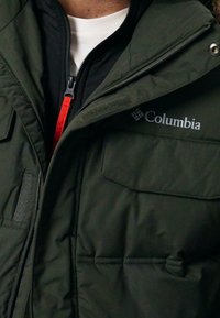 Tamnozelena Columbia jakna s patentnim zatvaranjem, s jastučenom teksturom i istaknutim logom. Ima crveni povlačak za patentni zatvarac i dva prednja džepa.