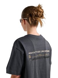 Kobieta w okularach przeciwsłonecznych i z niechlujnym kokiem, ubrana w ciemnoszarą koszulkę z napisem „HIGH ALTITUDE LOW OPENING” na plecach.