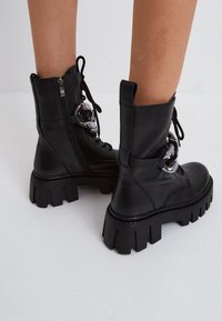 Bottes bottines en cuir noir avec une semelle épaisse, dotées d'une fermeture éclair et d'un laçage. Accents métalliques argentés remarquables et finition texturée.