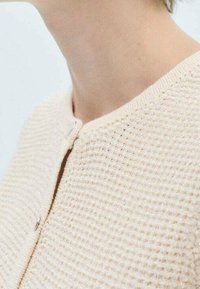 Jasno beżowy kardigan o fakturowanej strukturze z plecionym wzorem, okrągłym dekoltem i małym srebrnym zapięciem z przodu. Prosty design, neutralny kolor.