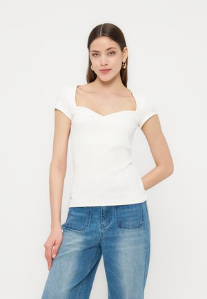 Kvinde med langt brunt hår iført gyldne øreringe i hoops, en hvid kortærmet tætsiddende top med firkantet udskæring og blå denimjeans, stående med hånden bag ryggen.