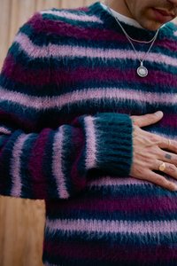 Striped knit sweater featuring pink, purple, and teal hues, with a soft, fuzzy texture and ribbed cuffs. Accented by simple jewelry.