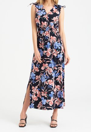 Žena v černých maxi šatech bez rukávů s růžovo-modrým květinovým potiskem, s bočním rozparkem a černými sandály na podpatku.
