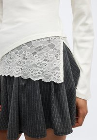 Λευκό μακρυμάνικο τοπ φορεμένο πάνω από λευκή φλοράλ δαντέλα, σε συνδυασμό με σκούρο γκρι πλισέ φούστα με ρίγες, εμφανιζόμενο από τη μέση έως το μέσο του μηρού.