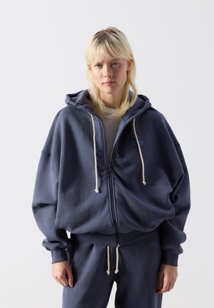 Persona bionda che indossa una felpa con cappuccio blu navy oversize con cerniera e coulisse, abbinata a pantaloni della tuta coordinati, in piedi su uno sfondo neutro.