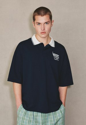 Junge Person mit kurzen Haaren, die ein marineblaues Poloshirt mit weißem Kragen und grüne karierte Hose trägt, steht mit den Händen in den Taschen.