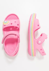 Vaaleanpunaiset lasten sandaalit, jotka on valmistettu kevyestä kumista. Niissä on sateenkaariaskartelu, säädettävä hihna ja värikkäät raidoitetut pohjat.