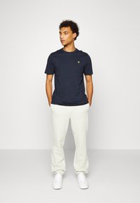 T-shirt blu navy a maniche corte con logo giallo sul petto, abbinata a pantaloni della tuta grigi chiari e sneakers bianche. Il modello è in piedi.