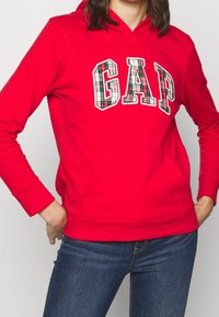 Červená mikina s kapucí s výrazným logem "GAP" a kostkovanými detaily. Látka má jemný povrch a obsahuje kapsu vpředu.