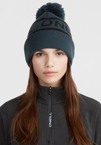Knit beanie in dark teal with a black logo band and a pom-pom on top; person wearing a black zip-up fleece jacket.