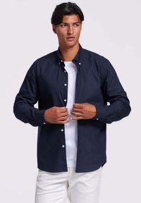 Jonge man die een marineblauwe button-up shirt over een wit T-shirt aanpast, gekleed in witte broek, staand tegen een lichte achtergrond.