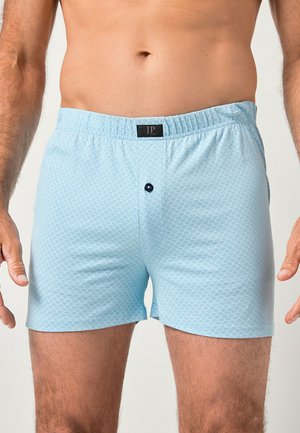 Mężczyzna w jasnoniebieskich bokserkach w wzory z zapinaniem na guziki i elastyczną talią, stojący na jednolitym tle.